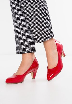 Jambes portant un pantalon pied-de-poule noir et blanc associé à des chaussures rouges brillantes à talon bas sur fond blanc.