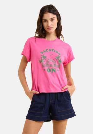 Rosa Baumwoll-T-Shirt mit kurzen Ärmeln, das mit grüner "VACATION ON"-Schrift und Grafiken von Palmen versehen ist. Kombiniert mit dunkelblauen Denim-Shorts.