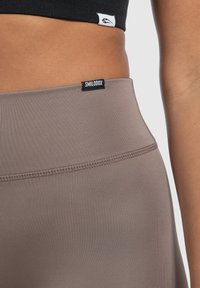 Leggings marroni a vita alta con texture liscia e dettagli di cucitura. Presentano un'etichetta nera con "SMILDOX" cucita sulla vita.