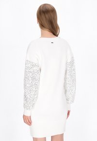 Rochie albă tricotată cu mâneci puff texturate și cu model. Mânecile au aplicații de paiete, iar marginea are un design cu côrdă. Partea din spate include un logo mic.