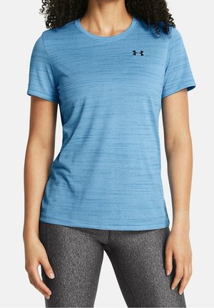 Kobieta ubrana w dopasowany niebieski krótki rękaw Under Armour oraz szare teksturowane legginsy, stoi z ramionami swobodnie opuszczonymi po bokach.