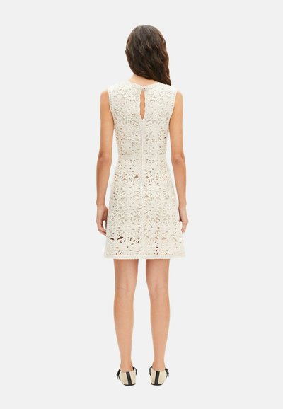 Mujer con vestido de encaje blanco sin mangas hasta la rodilla con patrones florales, cierre de botón en la parte trasera con abertura en forma de ojo de cerradura, y zapatos planos en blanco y negro.