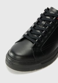 Black leather sneaker with textured upper, round toe, and black laces. Features a rubber sole and logo detail on the heel.
