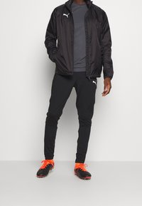 Black lightweight jacket, gray t-shirt, fitted black pants, and black soccer cleats with orange accents. Features Puma logos on clothing.