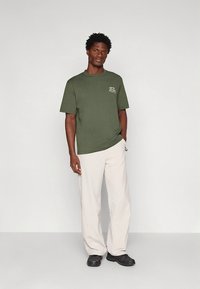 Olijfgroen katoenen t-shirt met een ronde hals en een klein wit motief op de borst, gecombineerd met lichtbeige broek en zwarte schoenen.