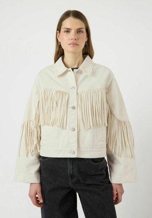 Kvinne iført en kremfarget jakke med knapper og frynser over brystet og ermene, kombinert med høytlivs svarte jeans.