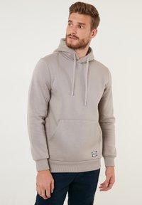 Sudadera gris de tejido suave con un bolsillo tipo canguro, capucha con cordón y puños de canalé en la parte inferior. Presenta una pequeña etiqueta de la marca en el bolsillo.