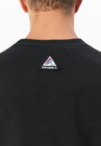 Närbild av en persons nacke och övre rygg som bär en svart skjorta med en triangulär O'Neill-logotyp nära kragen.