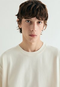 Joven con cabello corto y ondulado de color castaño, expresión neutral, vestido con una camiseta blanca lisa de cuello redondo sobre un fondo gris claro.