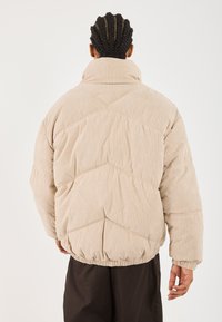 Doudoune en velours côtelé beige avec un design matelassé horizontal. Le col montant et l'ourlet élastiqué offrent une coupe décontractée.