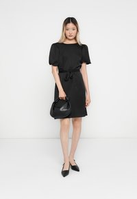 Robe noire avec manches bouffantes, taille cintrée avec ceinture, et longueur au-dessus du genou. Le mannequin tient un petit sac noir et porte des escarpins noirs à bout pointu.