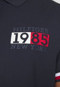 Marineblauwe polo met geborduurd ontwerp met "HILFIGER," "1985," in witte en rode secties, en "NEW YORK" in zwart, textuurstof.