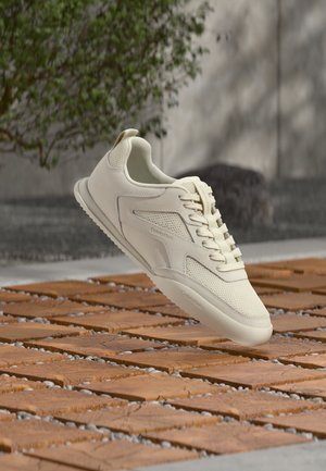 Baskets couleur crème avec lacets, flottant au-dessus d'une surface en briques et pierres aux motifs, avec des plantes floues et un mur en arrière-plan.