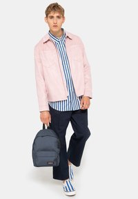 Rosa mockajacka över en blå- och vitrandig skjorta, marinblå byxor och blårandiga skor. Håller en grå ryggsäck med en framficka.