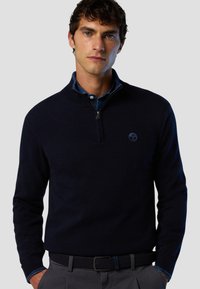 Marineblauer Pullover mit einem Halb-Reißverschluss-Kragen, aus weichem Stoff gefertigt. Verfügt über ein Logo auf der Brust, kombiniert mit einem hellblauen Hemd mit Kragen darunter.