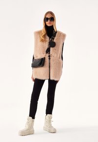 Gilet in pelliccia sintetica rosa chiaro, abbinato a leggings neri e stivali crema. Accessoriato con una piccola borsa a tracolla nera e occhiali da sole grandi.