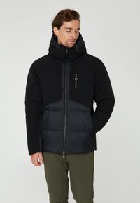 Svart puffjacka med dragkedja framtill, kviltad nederdel och hög krage. Har horisontella och vertikala paneler samt en logotyp.