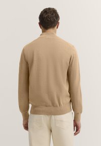 Beige Strickjacke mit geripptem Saum und Bündchen, die eine lässige Passform bietet. Der Stoff wirkt weich mit einer glatten Textur.
