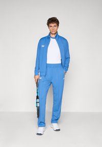 Blå sportsjakke og bukser i letvægtstof, med en hvid t-shirt under, som holder en tennisketsjer. Enkle logo accenter.