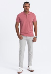 Polo rouge avec une finition texturée, présentant une poche poitrine. Associé à un pantalon slim gris clair et des baskets blanches avec des accents marron.