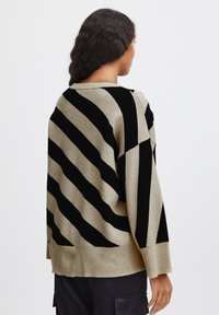 Pull en tricot beige avec de larges rayures noires diagonales, une coupe décontractée, un ourlet côtelé et de larges manches. Finition texturée pour un intérêt visuel accru.
