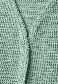 Tissu texturé vert clair avec un motif en maille gaufrée, présentant un design à l'avant drapé et des bords légèrement festonnés le long de l'encolure.