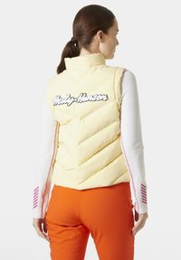 Světle žlutá péřová vesta s chevronovým prošíváním, vyšívané logo "Helly Hansen" v černé barvě, nošena přes bílé tričko s dlouhým rukávem a oranžové kalhoty.