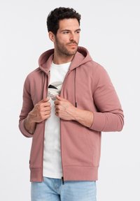 Sweatshirt à capuche zippé en tissu rose pâle avec une texture lisse, présentant une poche kangourou et des poignets côtelés. Porté par-dessus un t-shirt blanc.