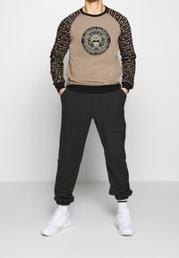 Felpa beige con maniche a motivo nero, con una grafica centrale di un leone. Abbinata a pantaloni jogger scuri e sneakers bianche.
