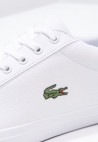 Gros plan sur une basket en toile blanche avec des lacets blancs et un logo crocodile vert cousu sur le côté au-dessus de la semelle blanche.