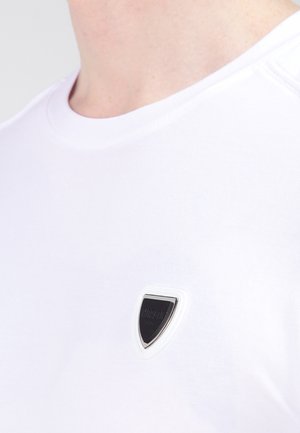 T-shirt en coton blanc avec un col rond et un petit logo en forme de bouclier noir et blanc sur le côté gauche de la poitrine. Texture lisse, design simple.