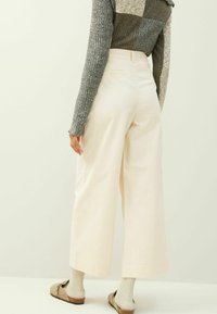 Brede broek in crème stof met een gladde textuur en een getailleerde pasvorm, gecombineerd met beige schoenen en een lichtgrijs geribbeld trui.