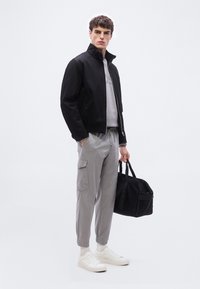 Νεαρός άνδρας στέκεται κρατώντας μαύρη τσάντα duffel, φορώντας μαύρο μπουφάν, γκρι παντελόνι cargo, λευκά αθλητικά παπούτσια και ανοιχτό γκρι φούτερ σε λευκό φόντο.