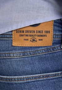 Nahaufnahme von blauen Denim-Jeans mit einem braunen Lederetikett der Marke, auf dem steht: „Denim Driven Seit 1989, hochwertige Kleidung gefertigt.“
