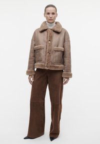 Bruine schapenvachtjack met een kraag, twee voorzakken en knoopsluiting. Gepaard met bruine wijde broek en puntige zwarte schoenen.