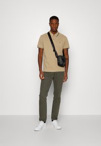 Polo shirt tan con dettagli verdi, pantaloni verde oliva e scarpe da ginnastica bianche, abbinati a una piccola borsa a tracolla nera. Sfondo semplice.