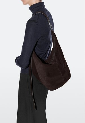 Frau in dunklem Rollkragenpullover und Rock trägt große dunkelbraune Veloursleder-Schultertasche mit Reißverschluss und langem Trageriemen.