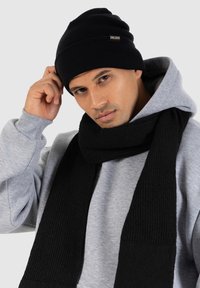 Beanie e sciarpa neri a coste realizzati in materiale di maglia testurizzata. Indossato con una felpa leggera grigia. Il beanie presenta un piccolo logo.