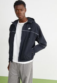 Mladić s kratkom kovrčavom kosom u crnoj Nike trenirci s patentnim zatvaračem preko bijele majice i svijetlosivih hlača, stoji s jednom rukom u džepu.