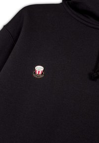 Sudadera negra con una pequeña insignia de esmalte en forma de cubo de palomitas de maíz con rayas rojas y el texto "Disney BSL" en el lado izquierdo del pecho.