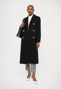 Donna che indossa un lungo cappotto nero doppiopetto, camicia bianca, pantaloni a quadri, tacchi bianchi, con una borsa blu navy a spalla, in piedi.