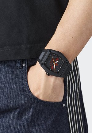 Črno uro s kvadratno obliko, teksturirano črno glavna plošča, rdeči detajli, oranžne kazalce in gumijast trak. Nosena na zapestju ob temno modrih jeansih.
