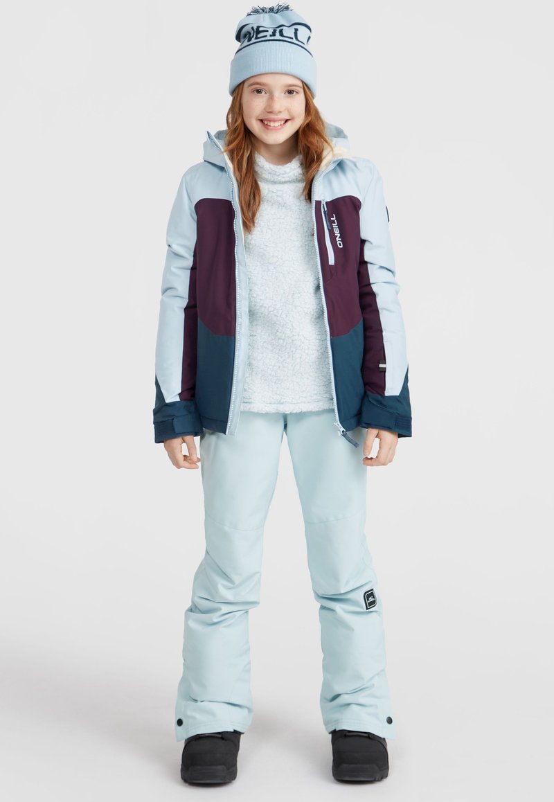 Veste d'hiver bleu clair avec des accents marine et bordeaux, associée à un pull en laine bouclée bleu clair et à un pantalon de ski. Des bottes de neige noires complètent la tenue.