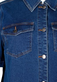 Jeans košulja plave nijanse, s dva džepa na prsima, metalnim gumbima i kontrastnim narančastim šavovima.