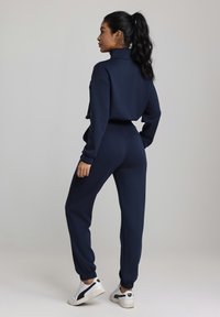 Marineblå joggedress med høy krage, ribbestrikkede mansjetter og elastisk linning. Kombinert med hvite og svarte joggesko. Glatt, elastisk stoff.