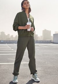 Olijfgroen athleisure set met een ritsjack en taps toelopende broek, gemaakt van zacht materiaal en met een relaxte pasvorm, gecombineerd met lichte sportschoenen.
