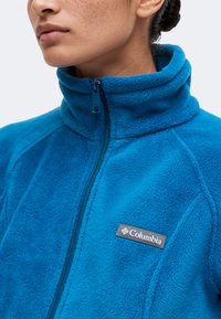 Γυναίκα φοράει έντονα μπλε fleece μπουφάν Columbia με ψηλό κολάρο, φερμουάρ ως επάνω, με το λογότυπο στο στήθος.