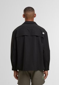 Mężczyzna z krótkimi włosami, ubrany w luźną czarną koszulę z długim rękawem i oliwkowe szorty, stojący tyłem na jednolitym szarym tle.