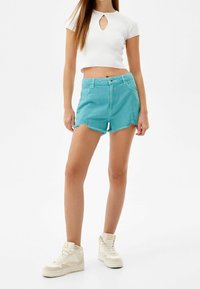 Shorts de mezclilla turquesa con dobladillos deshilachados, cintura alta y bolsillos delanteros, combinados con un crop top blanco acanalado con un corte en forma de ojo de cerradura.