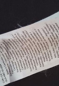 Valkoinen materiaalin hoito-ohje etiketti mustalla tekstillä, joka sisältää pesuohjeet ja materiaalitiedot. Sisältää useita kieliä ja symboleita.